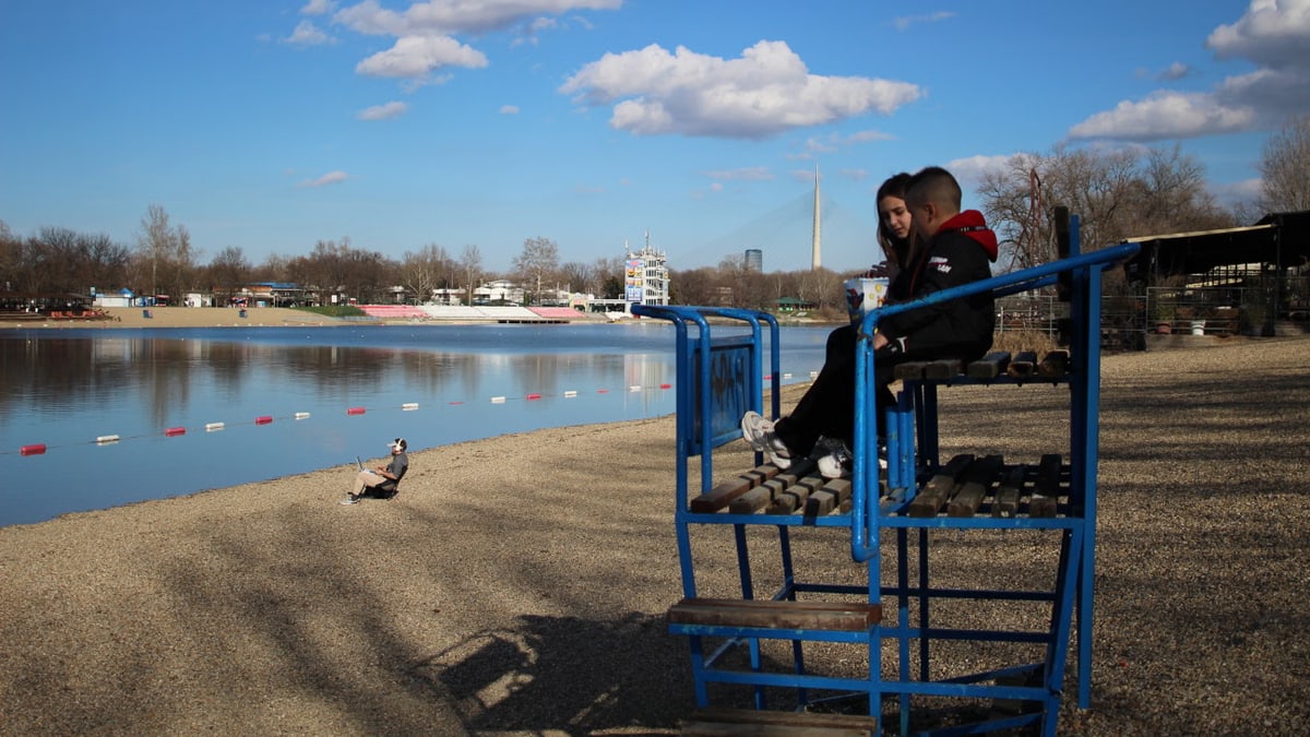 A young couple sits on a lifeguard chair by a serene river under a bright blue sky.